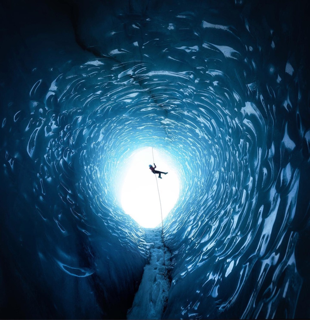 a lone figure silhouetted in the entrance of a blue ice cave, descending on a rope