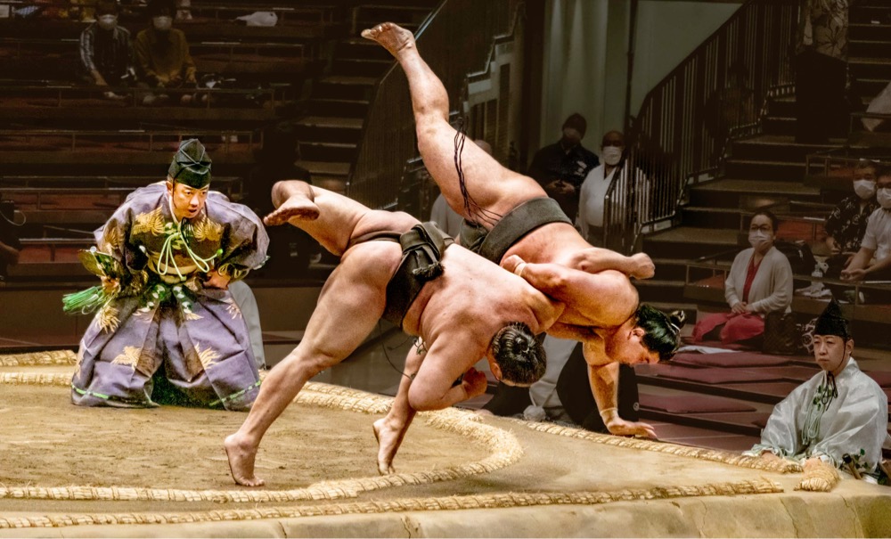 a sumo wrestler throws another wrestler out of the ring while the judge carefully watches